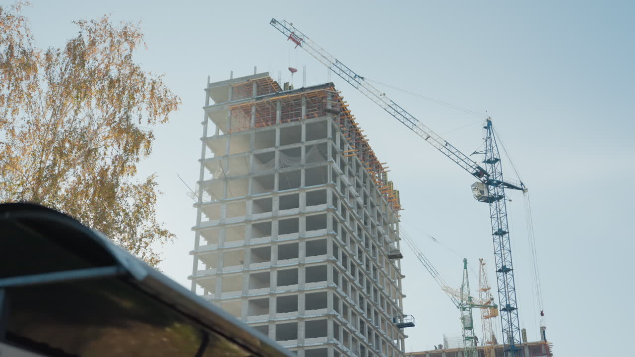 Torre di cemento illuminata dal sole con gru, betulle autunnali che incorniciano il cantiere, tettoia di una fermata dell'autobus in primo piano, solai scheletrici e travi a vista, calda retroilluminazione, lenta panoramica cinematografica che trasmette una calma urbana.