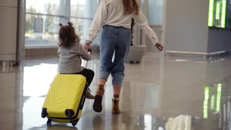 Airport-terminal,-young-unrecognizable-mother-riding-her-cute-daughter-on-a-small-yellow-suitcase.-Mom-with-yellow-suitcase-and-daughter-are-having-fun-before-their-departure