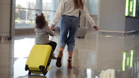 airport terminal, young unrecognizable mother riding her cute daughter on a small yellow suitcase. mom with yellow suitcase and daughter are having fun before their departure