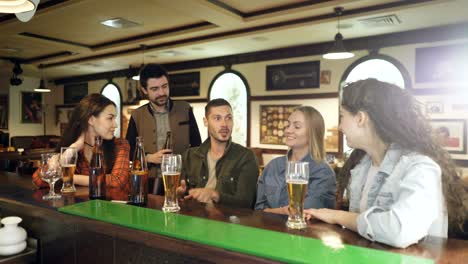 dark-haired bearded young man is telling a story and gesturing emotionally while meeting with his friends in pub at counter. his mates are listening to him carefully.