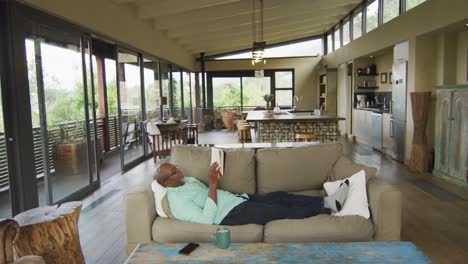 happy african american senior man relaxing in living room, lying on couch reading a book