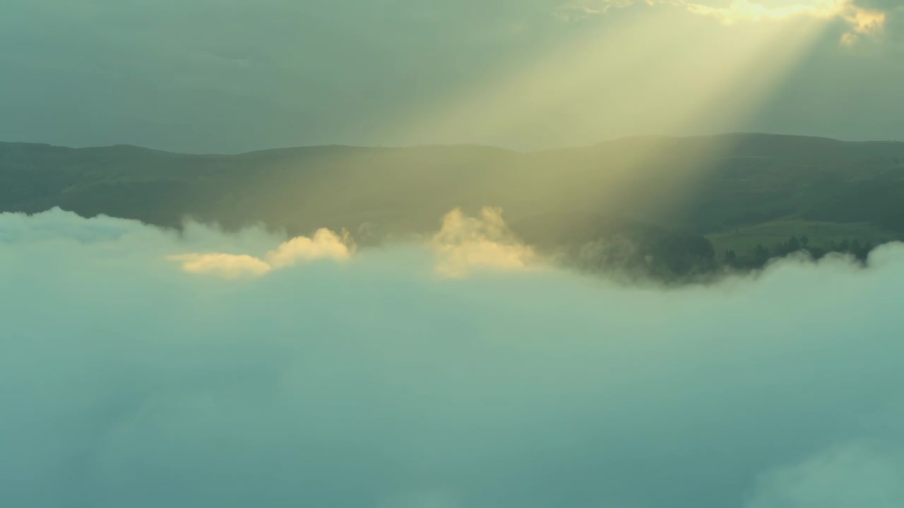 Piercing sunbeam breaking through cloud gap, illuminating low cloud bank and puffs from hilltop