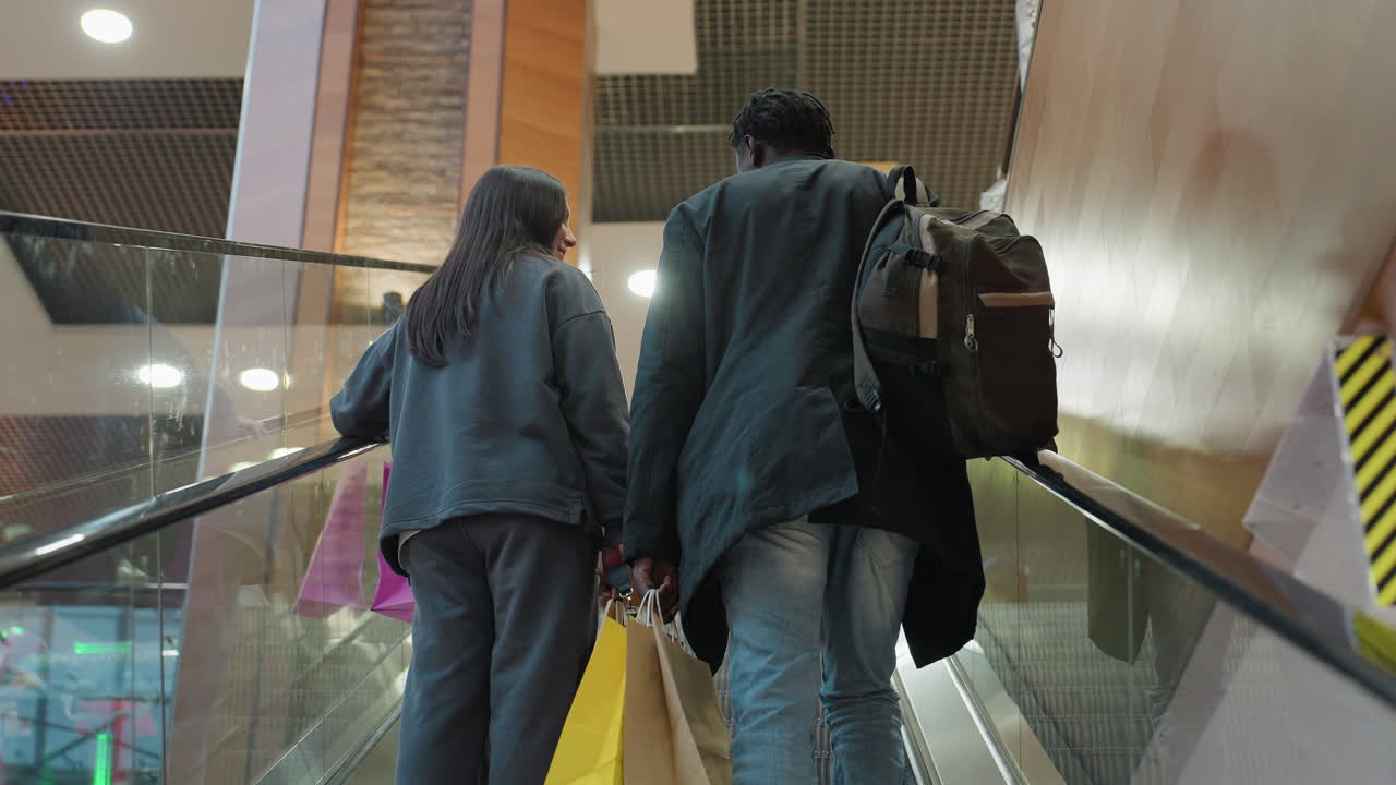 Rear view of two young shoppers holding colorful bags while riding ascending escalator inside stylish commercial shopping center with glass walls, warm lights, and urban interior design