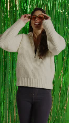 video vertical de una mujer celebrando el día de san patricio de pie frente a una cortina de oropel verde con prop shamrock deely boppers y gafas