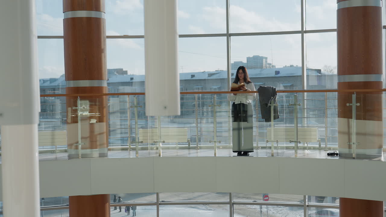 Young student in white shirt stands near glass railing inside mall gallery, reading book attentively as sunlight filters through large windows with visible reflection of parked cars outside