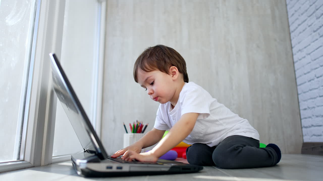 Positive baby boy sits on his knees near the laptop. Curious toddler pressing the keys on the keyboard. Low angle view.