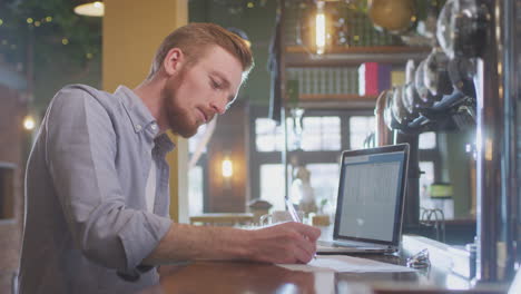 Propietario-Masculino-O-Gerente-De-Bar-Trabajando-En-Una-Computadora-Portátil-En-El-Mostrador-Haciendo-Cuentas