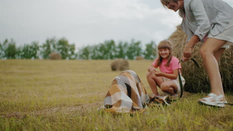 joyful moment as dog playfully covered with blanket, owner laughs while opening it, daughter squats nearby enjoying the fun scene, warm outdoor setting with hay bales, countryside ambiance