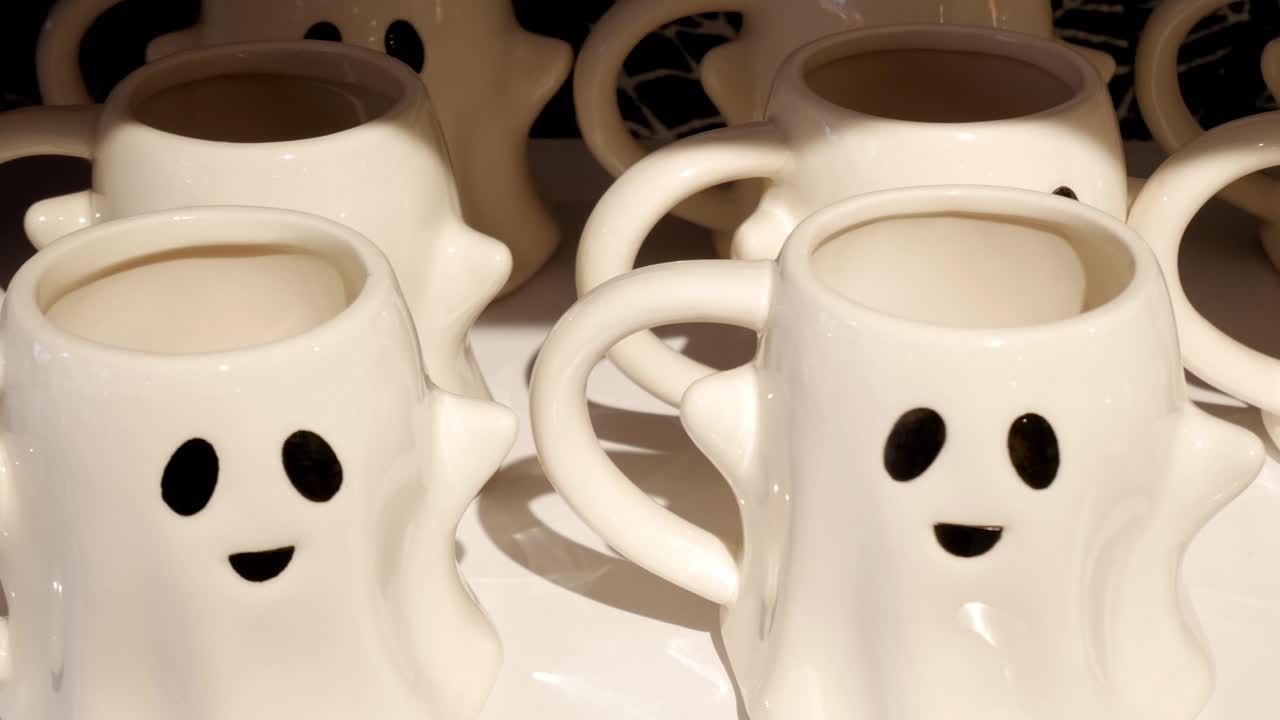 Geisterförmige Tassen für Halloween, reihenweise ausgestellt. Freundlich aussehendes Gesicht mit schwarzen Augen und Mund. Hat auch geformte Arme an den Seiten