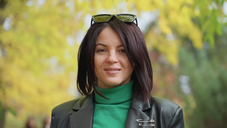 Donna con giacca di pelle che cammina in un parco autunnale illuminato dal sole, dolcevita verde e occhiali da sole, sorriso sicuro, profondità di campo ridotta, fogliame giallo, ritratto di moda casual, sfondo bokeh
