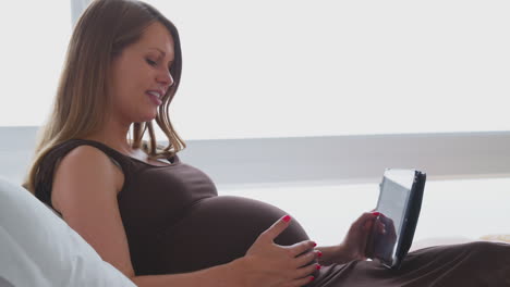 Schwangere-Frau-Entspannt-Sich-Zu-Hause-Im-Bett-Und-Schaut-Sich-Ein-Digitales-Tablet-An,-Das-Den-Bauch-Hält