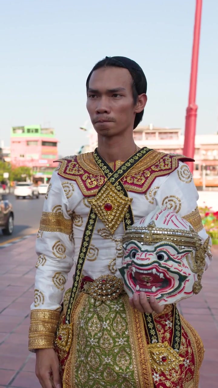 Seorang penari pria Thailand dengan kostum tradisional berhias berdiri di luar ruangan memegang topeng Hanuman.