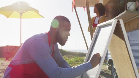 African-american-man-wearing-apron-writing-on-food-menu-slate-board-of-the-food-truck