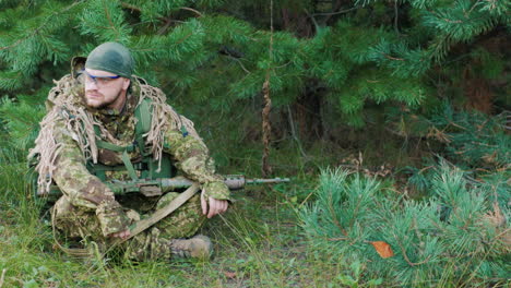 el joven de uniforme con un arma en las manos está sentado en el bosque descansando mirando alrededor