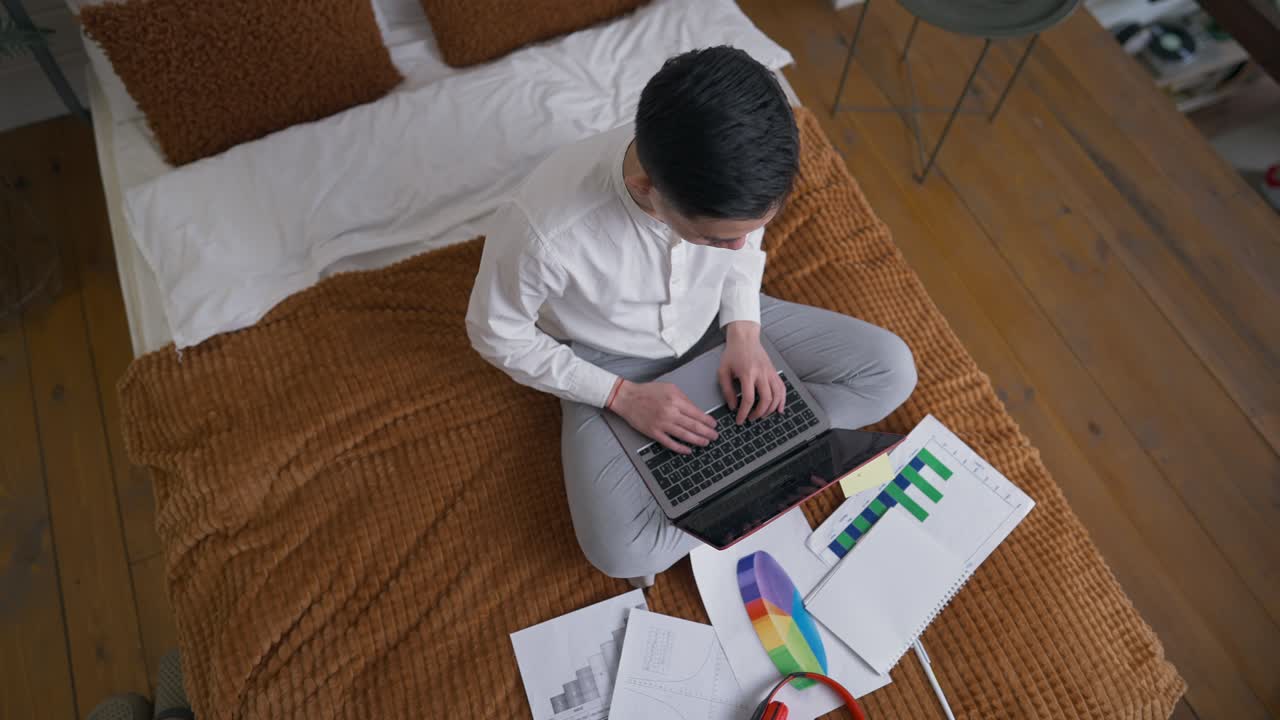 Top view of concentrated Asian young man typing on laptop keyboard sitting on bed at home office. Wide shot of expert professional analyst messaging online in bedroom. New normal coronavirus lifestyle.