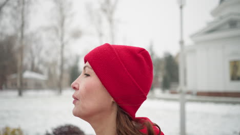 close up side view of tired athlete jogging along snowy park pathway in red beanie and hoodie, with frosty winter trees, and distant snow-dusted resident and white building in the background