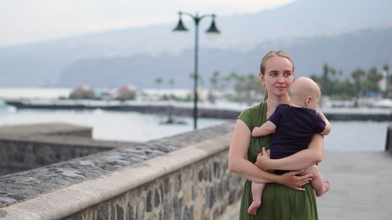 A young mother and her baby son take a walk on an ancient European square near the ocean, where they enjoy the waves and exchange heartfelt smiles