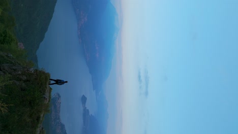un excursionista solitario está de pie en el borde de un acantilado, mirando hacia una impresionante vista panorámica de un lago sereno rodeado de montañas bajo un cielo azul claro, capturando un momento de asombro y reflexión