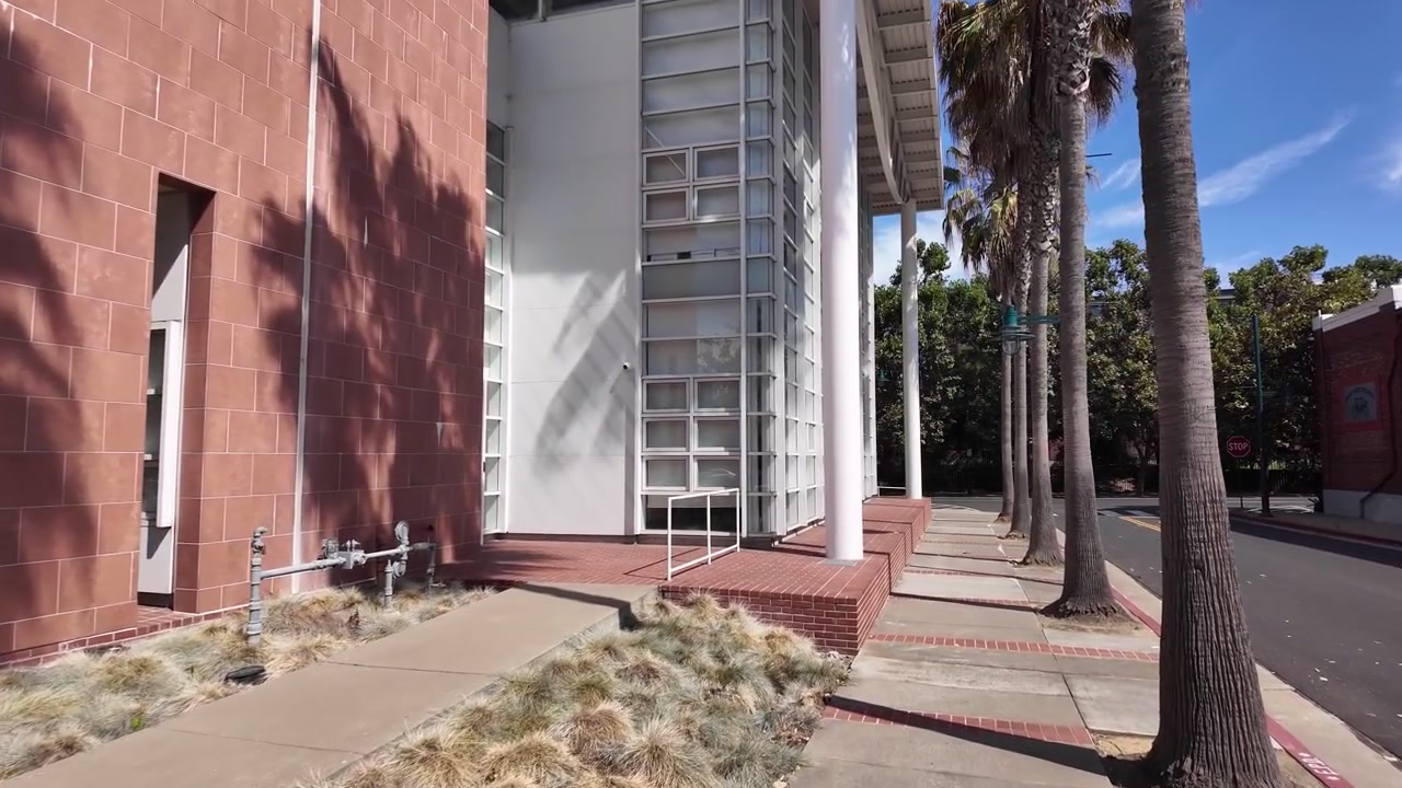 Una toma media captura la fachada iluminada por el sol del Centro Cívico de Emeryville en California, con modernas columnas blancas, ladrillo rojo, grandes ventanales de cuadrícula y aceras bordeadas de palmeras bajo cielos azules.