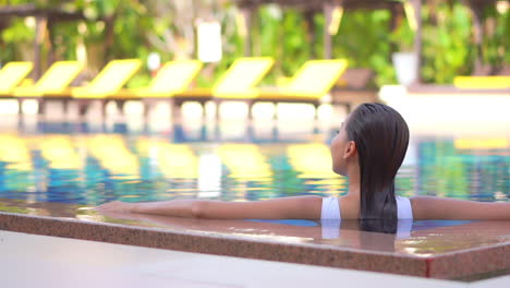 Back-of-American-woman-with-wet-hair-leaning-on-pool-border-inside-swimming-pool,-template-copy-space