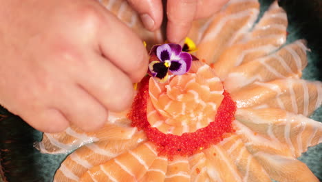 adding colorful flowers as decoration to a platter of delicious salmon sashimi with roe - close up