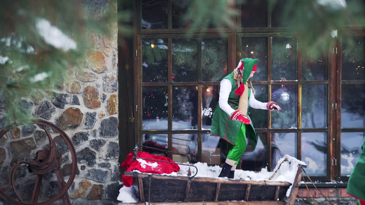 Joyful elves playing in snow. Two cheerful fairy characters in green suits having great time together near the big window outdoors. Christmas time