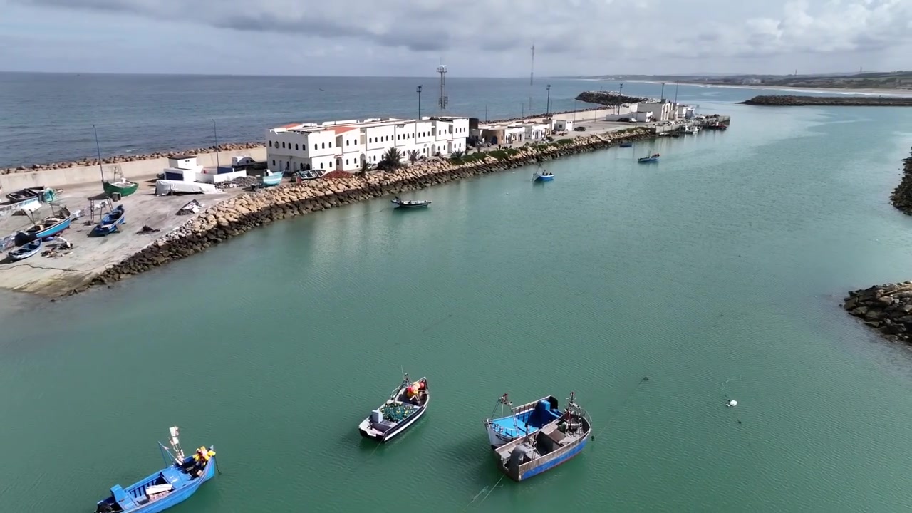 Vista aerea di piccole barche da pesca che galleggiano all'interno del porto di Asilah in Marocco, con acqua calma e protetta, strutture portuali operative e la costa atlantica sullo sfondo.