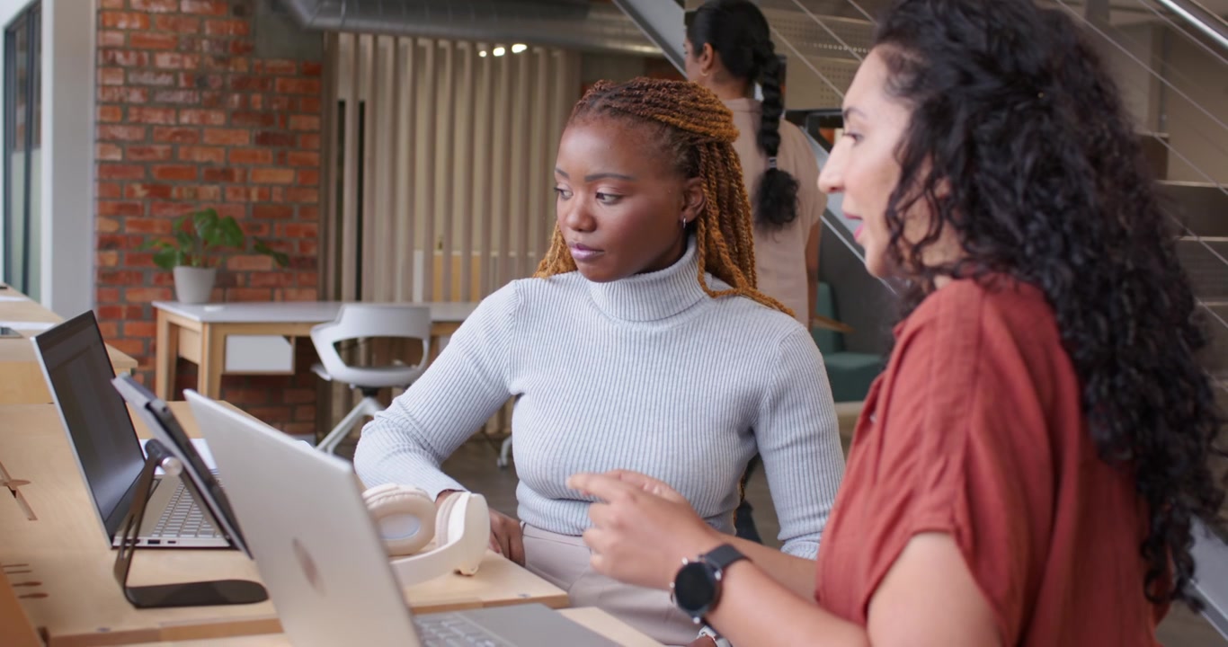 Female coworkers at desk clarifying, turtleneck woman raising hand, pointing at laptops and headset