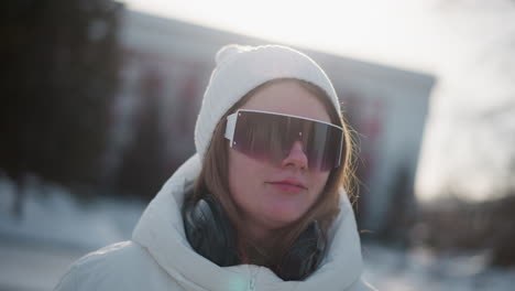 close up of creative young woman donning tinted shades and headphones while joyful swaying to music under bright winter sky wind tousles hair rosy cheeks and manicured nails catching sunlight