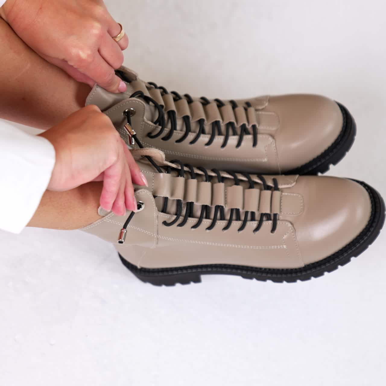 Female model sitting at studio holding by the tongues of stylish boots on her feet. Woman demonstrating fashionable modern footwear with black laces and black soles. Top view. White backdrop