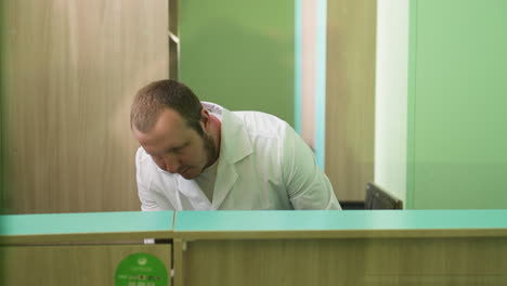 a close view of a lab technician in a white coat leaning forward, searching for something around with a stand shelf behind him