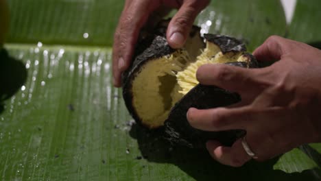 Fruta-Del-Pan-Asada-A-Fuego-Abierto,-Hombre-Rompiendo-Fruta-Del-Pan-Asada-En-Una-Hoja-De-Plátano-Con-La-Mano-Y-Abriéndola,-Comida-Tradicional-De-Seychelles,-Mahe,-Seychelles-4k-24fps-007