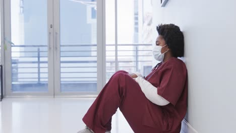 Tired-african-american-female-doctor-wearing-face-mask-sitting-in-hospital-corridor,-slow-motion