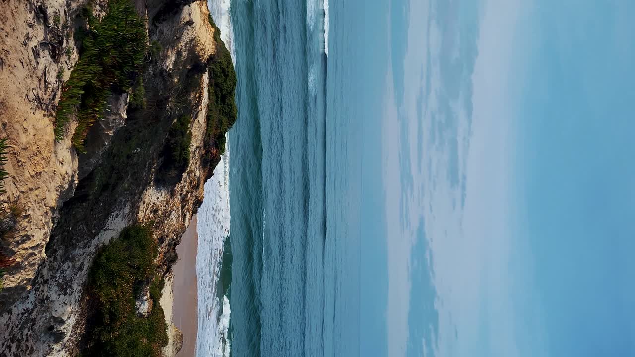 Zambujeira do Mar ved kysten med havbølger, klipper og sandklitter dækket af grøn vegetation, røde blade fra hottentotfigen, solrig dag, klar blå himmel