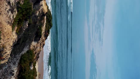 Zambujeira-do-Mar-over-the-sea-shore-with-ocean-waves,-cliffs-and-sand-dunes-covered-by-green-vegetation-red-leaves-of-sour-fig,-sunny-day,-clear-blue-sky