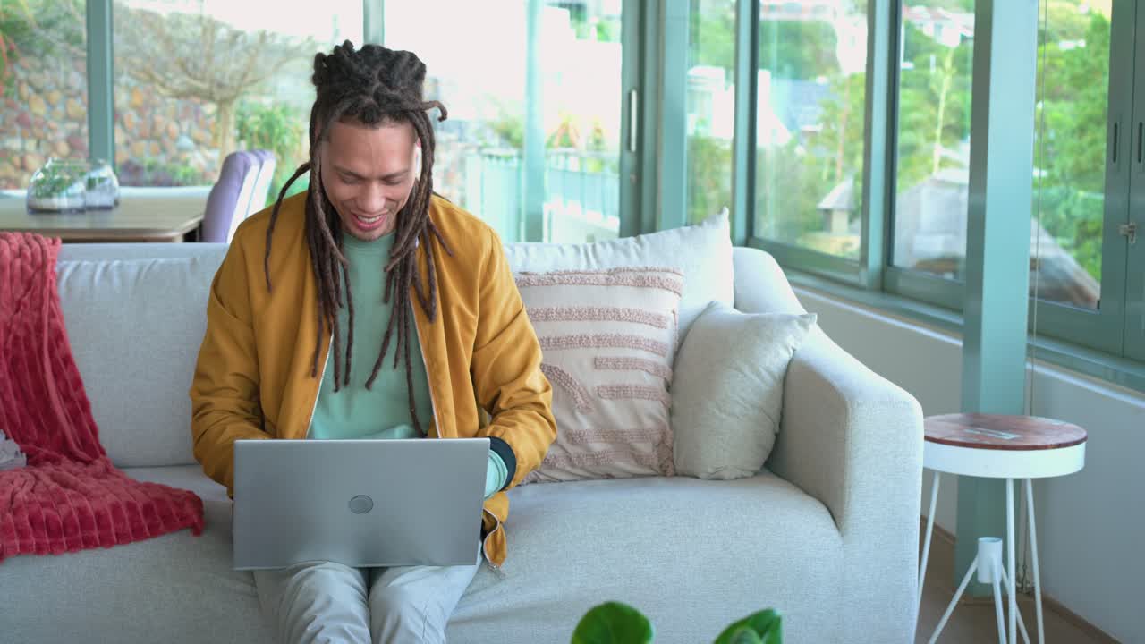 Man sitting on sofa opening laptop, typing, smiling at funny content, pausing on noise, refocusing