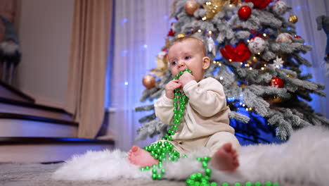 lovely little infant baby sitting at the christmas tree on the fluffy carpet. child plays with bright green string of beads and pulls them to his mouth. low angle view. vertical video.