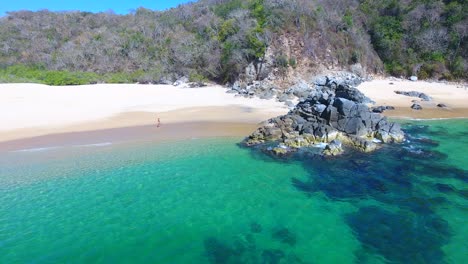 Niedrig-Schwebende-Antenne-Eines-Mädchens,-Das-Am-Weißen-Sandstrand-Zu-Einzigartigen-Felsformationen-In-Smaragdgrünem-Wasser-Geht