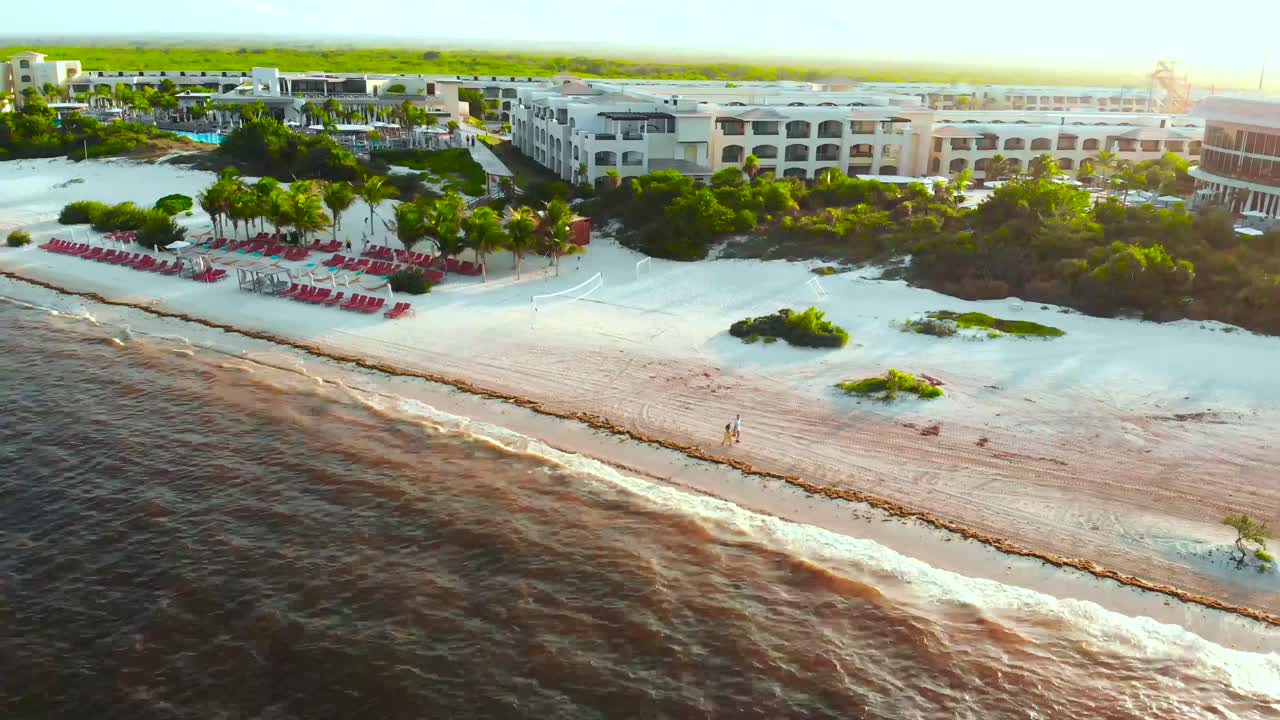A couple walks along the beach of a resort at sunset.