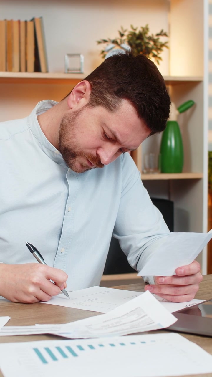 Giovane uomo al tavolo di casa che legge le pagine di un contratto e controlla i dettagli prima di firmare una lettera ufficiale.