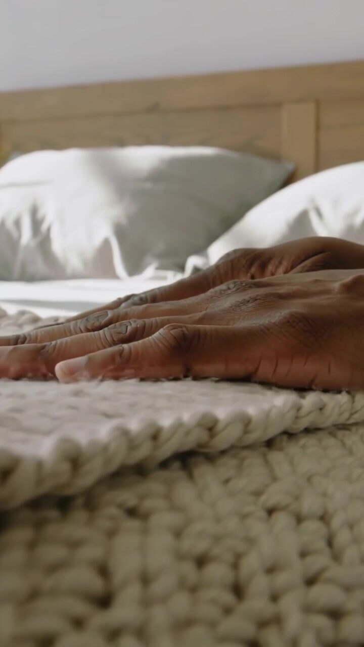 Video verticale: Mani scure che si posano lisciando un plaid a maglia sul letto, allineando il bordo ed esponendo il lenzuolo.