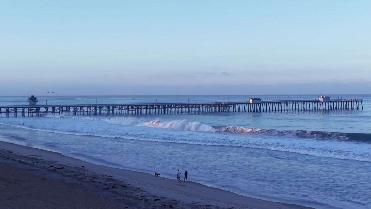 Un drone sorvola le coste della California meridionale. Due persone portano a spasso un cane sulla spiaggia alla luce del primo mattino, con l'iconico molo di San Clemente che si estende verso il mare come sfondo