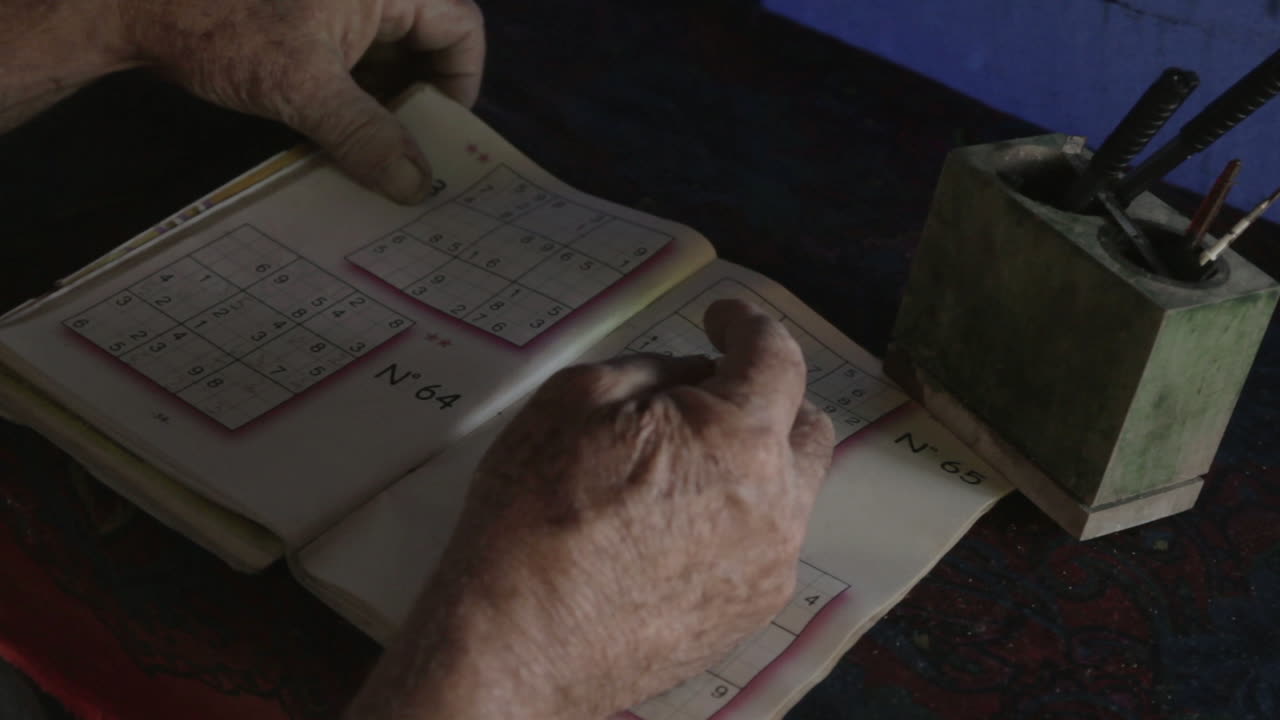 Worn hands move across a sudoku game in quiet focus, pencil in hand over an old desk tucked in Colombia’s jungle coast.
