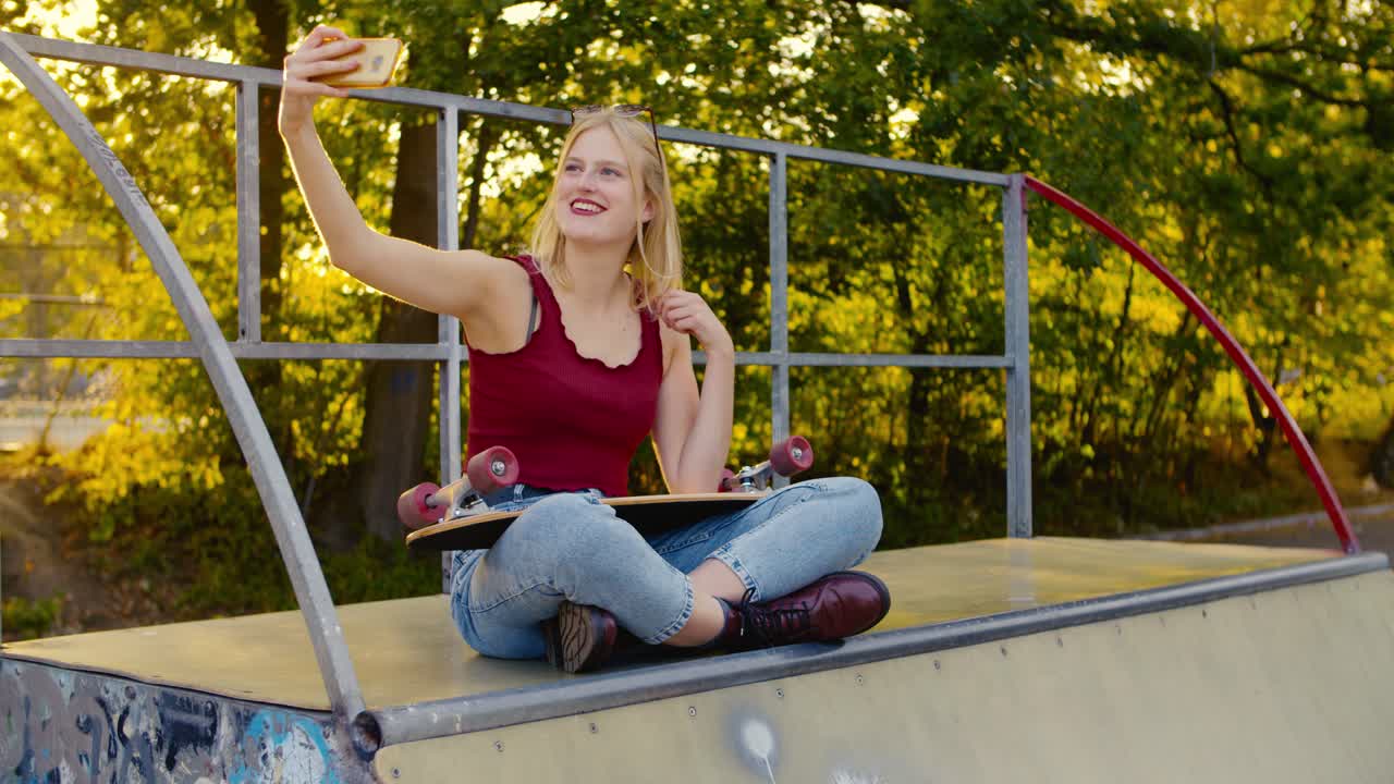 una patinadora feliz se está tomando una selfie sentada en un medio tubo al sol, con el sol detrás de su cabello rubio largo y una patineta en su regazo, riéndose de la cámara mientras posa, alta resolución