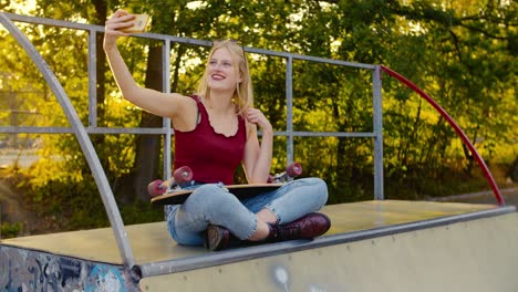 happy fit skater girl is taking a selfie sitting on a halfpipe in the sun, with the sun behind her blonde long hair and a skateboard on her lap, laughing at the camera while she poses, high resolution