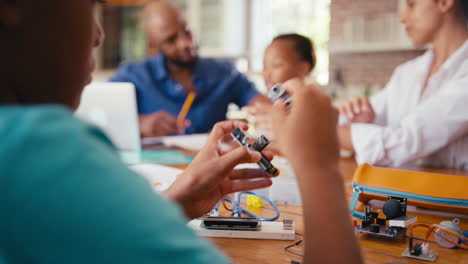 Familia-Alrededor-De-Una-Mesa-En-Casa-Usando-Una-Computadora-Portátil-Con-Padres-Ayudando-A-Los-Niños-Con-La-Tarea-De-Ciencias