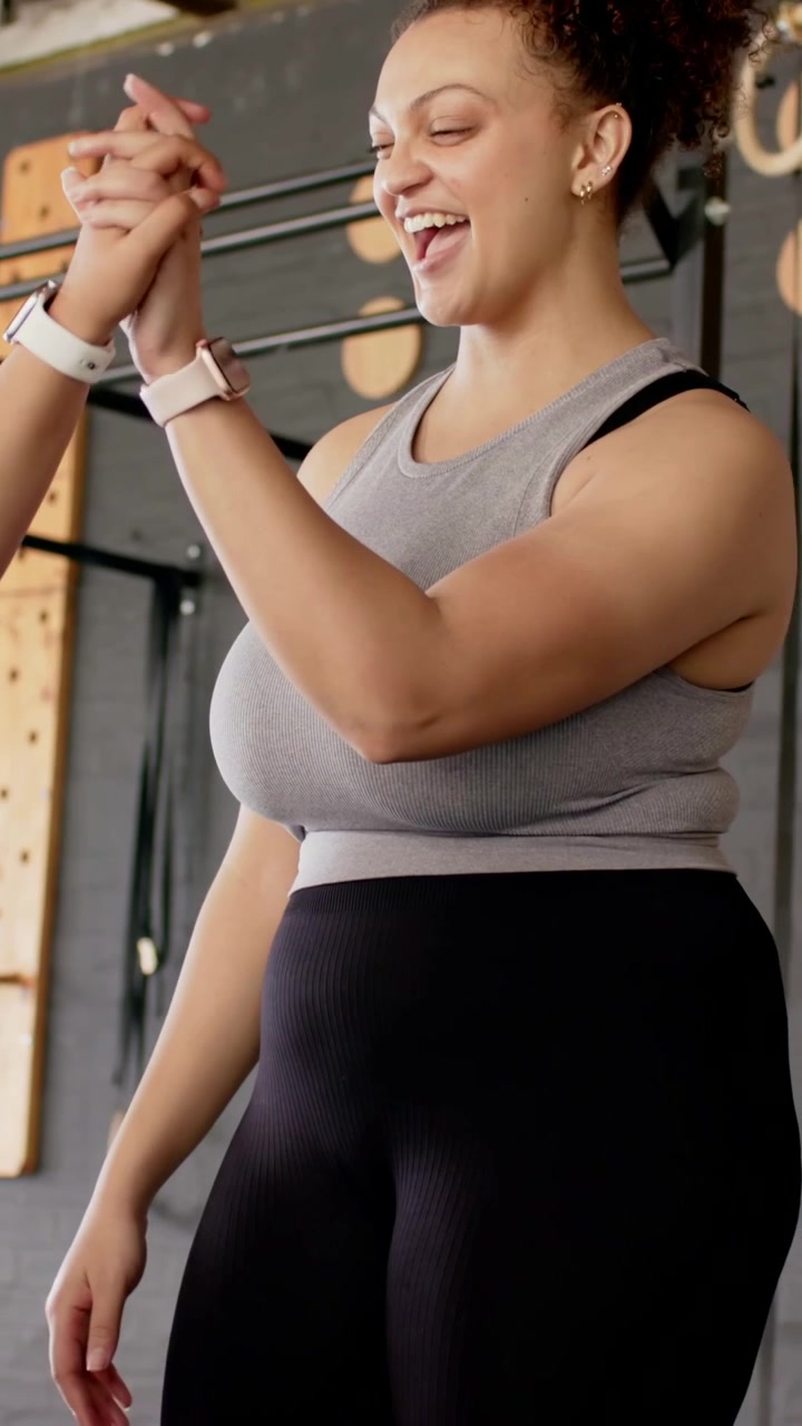 Video verticale: Donna inizia a fare curl con manubri, mentre l'assistente in maglietta grigia applaude e si stringe le mani in palestra.