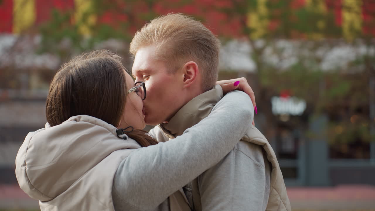 Lovers embrace affectionately as wife in glasses kisses her husband warmly while hugging close, dressed in matching outfits with vibrant building and passerby in background