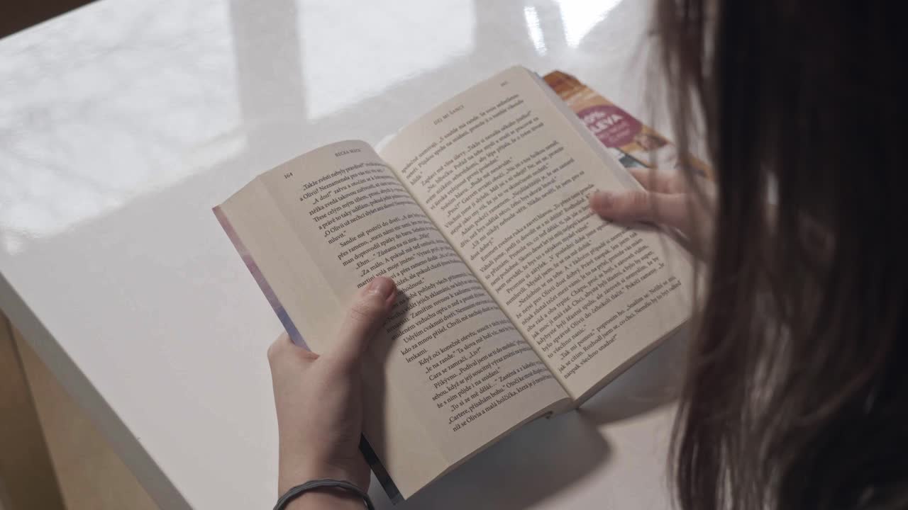 Inquadratura da dietro le spalle di una giovane donna che legge un libro cartaceo, usando un gesto di scorrimento del dito come su uno schermo di smartphone. Ideale per temi di psicologia sociale, abitudini, dipendenza dai media digitali o alfabetizzazione.
