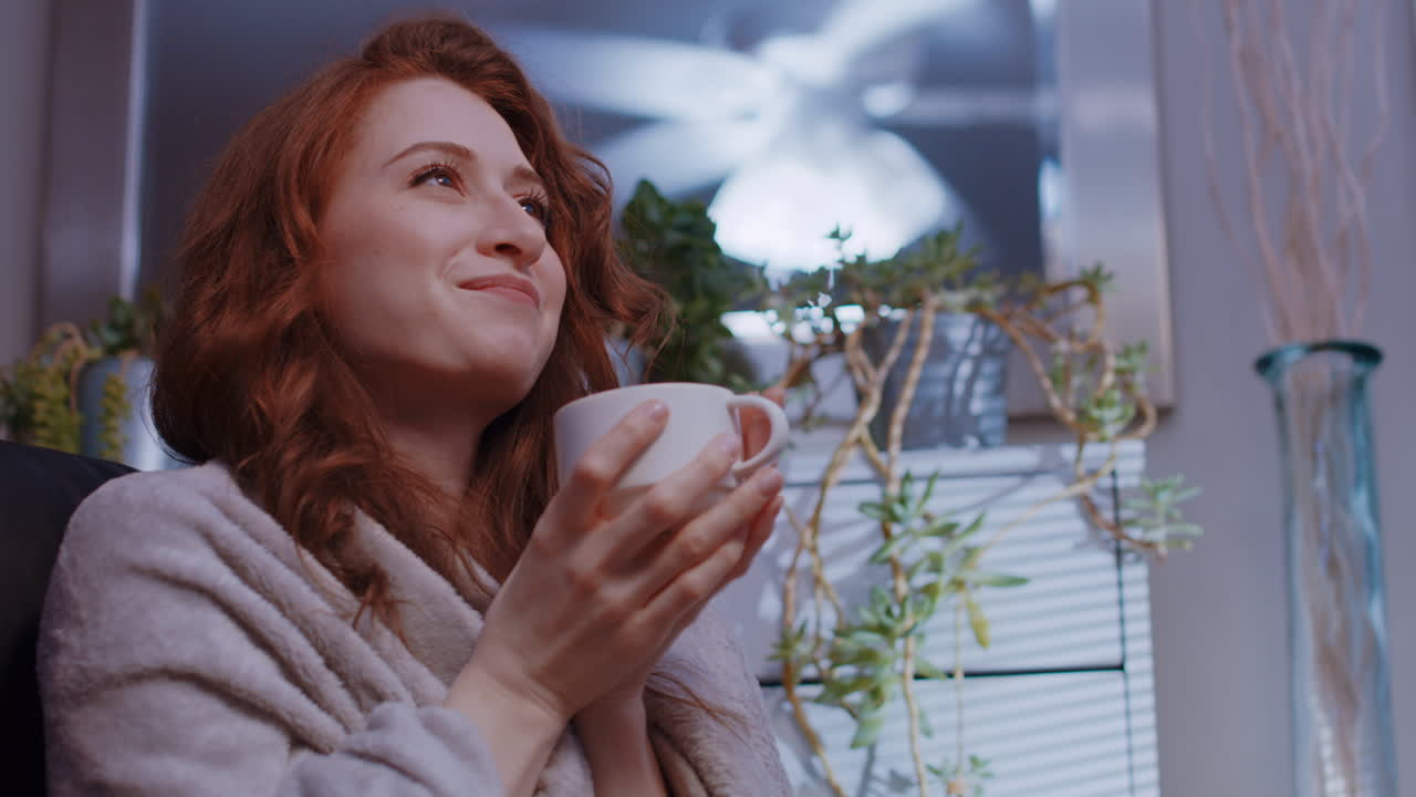 젊은 백인 여성이 집에서 편안하게 담요로 앉아 커피나 차를 마시고 행복합니다.